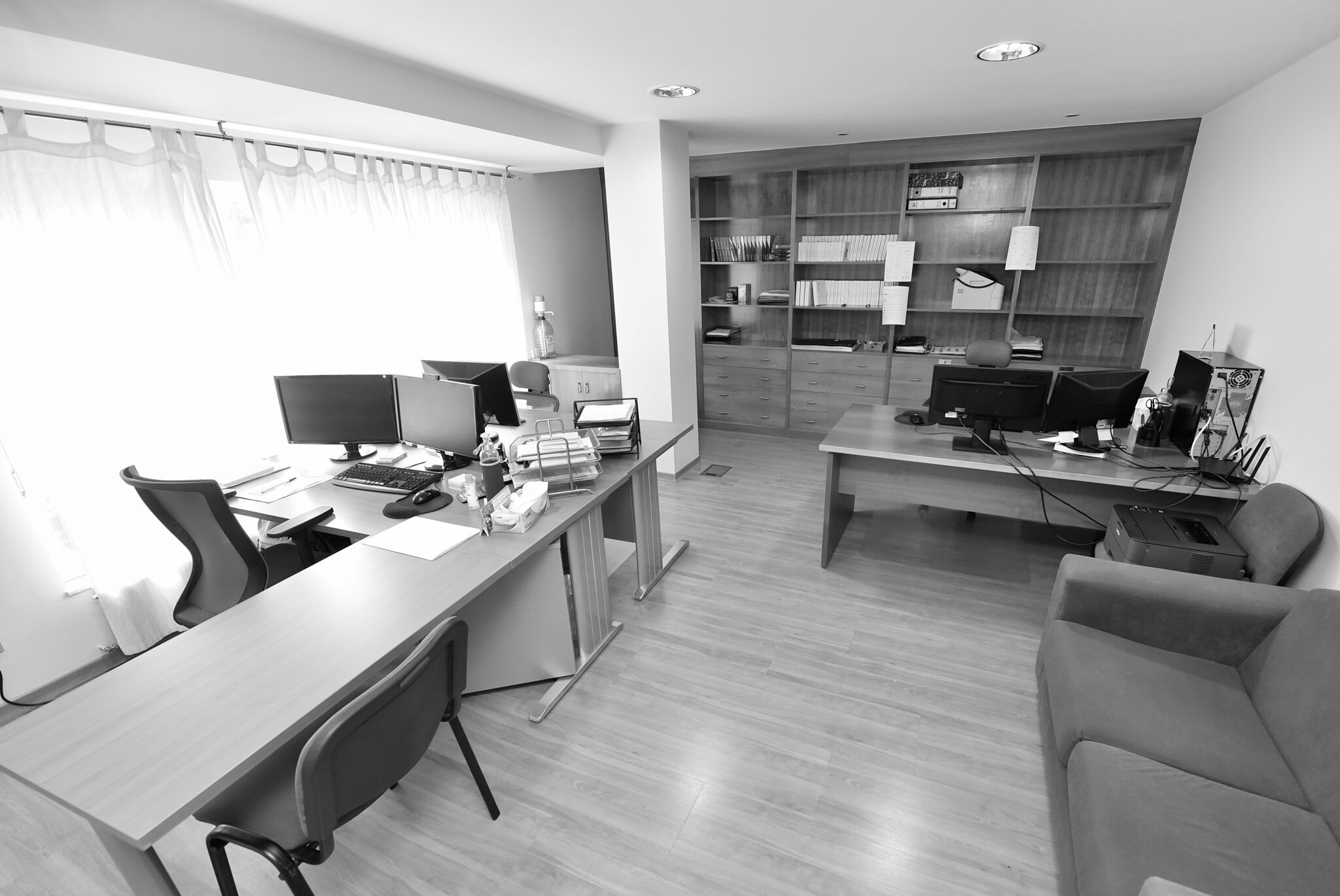 Oficina de Lugo con varios escritorios, ordenadores, estantería de madera y sillón junto a la mesa.