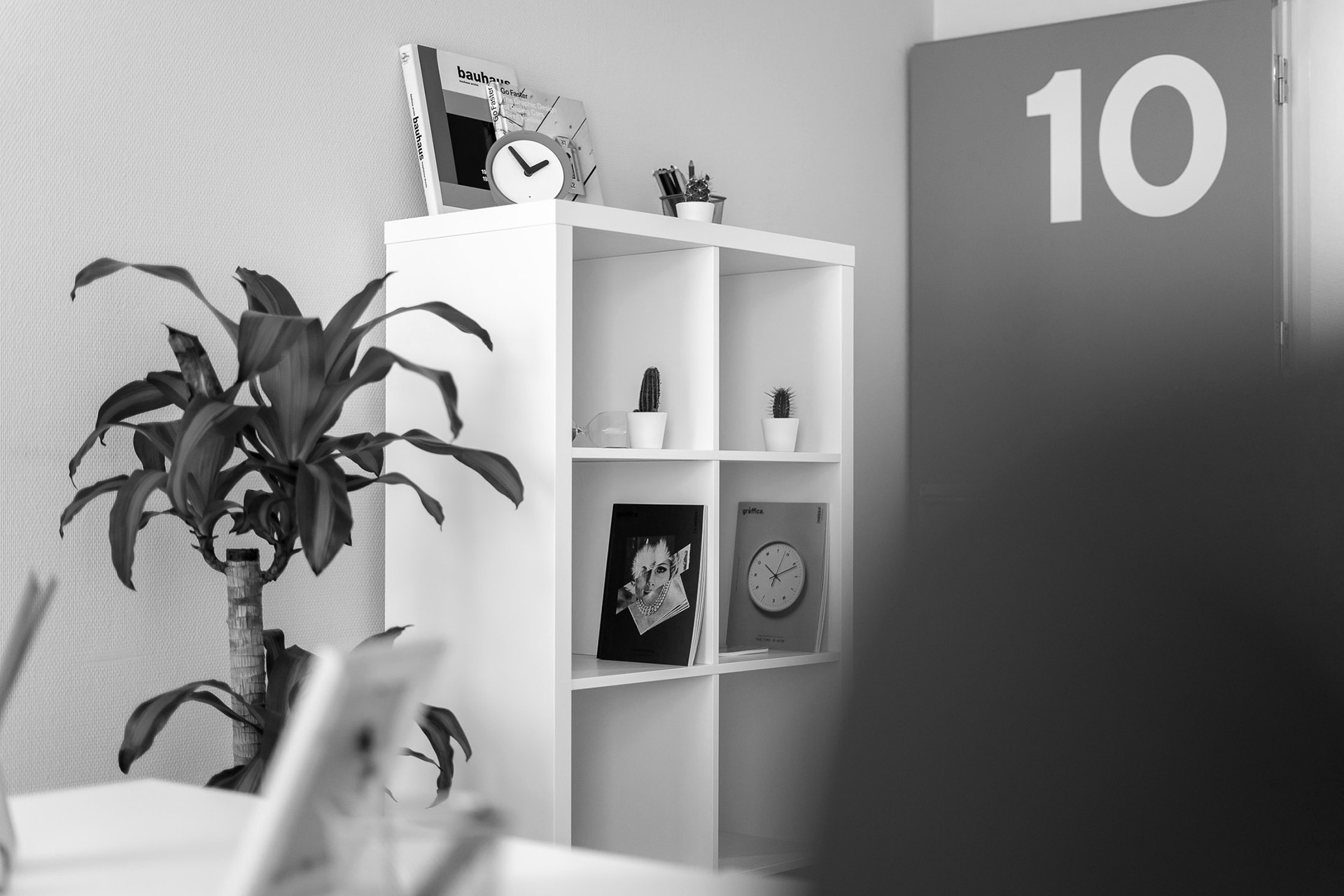 Entrada del despacho 10 de A Coruña con estantería blanca, planta y objetos decorativos.